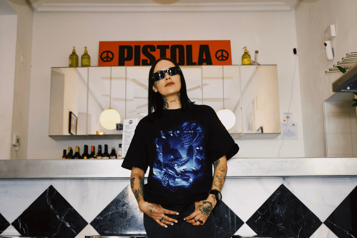 A woman with tattoos and sunglasses standing in front of a bar in a confident pose, wearing a t-shirt with an evil clown gripping a city and some sports cars