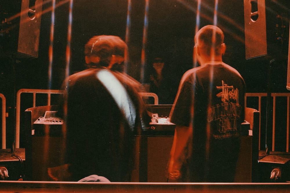 Two young men giving their back to the camera while DJing, with orange lighting and a dark environment